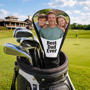May include: A golf club head cover featuring a custom photo of a parent and two children, with the text Best Dad Ever printed in black on a white background. The cover is positioned inside a black and white golf bag with several metal golf clubs.