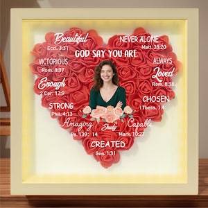 May include: A heart-shaped floral arrangement with a photo of a person in the center. The flowers are red and pink, and the arrangement is surrounded by a white frame. The frame has inspirational quotes and bible verses around the heart.