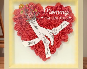 Può includere: Una cornice bianca con una disposizione a forma di cuore di rose rosse. All'interno del cuore, ci sono quattro mani che si tengono, con i nomi Riley, Avery, Katherine e Lily scritti sulle braccia. Il testo "Mommy We Love You" è scritto sopra le mani.