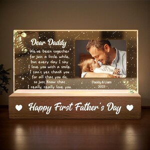 May include: A lighted Father's Day gift with a photo of a father and child. The acrylic panel has the words "Dear Daddy" and a loving message. The wooden base reads "Happy First Father's Day."