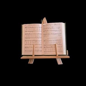 May include: An open book of sheet music rests on a small, light-colored wooden stand. The pages are filled with musical notation and text. The stand has a simple, A-frame design, and the background is black.