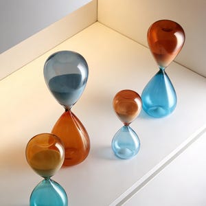 May include: Four glass hourglasses in shades of blue, orange, and amber are displayed on a white surface. The hourglasses vary in size and have contrasting coloured sand inside. A modern, decorative design.