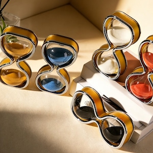 May include: A selection of hourglasses with yellow frames and clear glass. Each hourglass contains a different colour of sand: yellow, blue, white, red, and black. The hourglasses are displayed on a light-coloured surface.