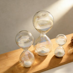 May include: Three clear glass hourglasses of different sizes, filled with white sand. The hourglasses have an iridescent finish, reflecting a rainbow of colours. They are arranged on a light brown wooden surface.