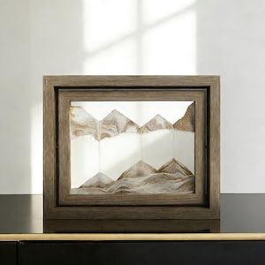 Puede incluir: Una pieza decorativa de arte de arena en un marco de madera. El arte de arena presenta un diseño de paisaje montañoso con tonos marrones y blancos. El marco es de forma cuadrada con un acabado de madera oscura, agregando un toque rústico.