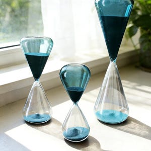 May include: Three glass hourglasses of varying sizes. Each hourglass has a clear, conical base and a teal-colored top filled with dark blue sand. The sand is flowing from the top to the bottom. The hourglasses are displayed on a white surface.