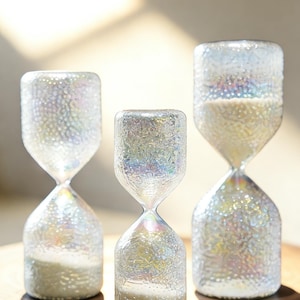 May include: Three iridescent glass hourglasses with white sand. The hourglasses have a textured, bubble-like surface and a classic shape. They are displayed on a wooden surface. The sunlight casts shadows across the scene.