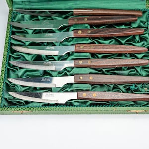 Puede incluir: Un juego de seis cuchillos de carne con hojas de acero inoxidable y mangos de madera oscura, presentados en una caja forrada de terciopelo verde. Cada hoja está marcada con el texto "St. Regis Stainless Japan".