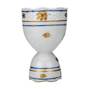 May include: White porcelain egg cup with gold and blue decorative accents. The egg cup features a basket weave pattern, gold floral designs, and blue stripes. The base and rim have gold trim. A classic design for serving eggs.