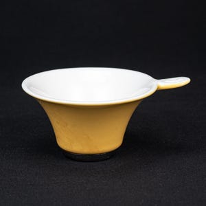 Puede incluir: Un pequeño embudo de cerámica de boca ancha con un exterior liso de color amarillo mostaza y un interior blanco limpio. El embudo presenta un asa pequeña y redondeada que se extiende desde el borde y una base de tono metálico oscuro.