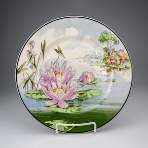 May include: A round ceramic plate featuring a hand-painted scene of pink water lilies floating on a pond with green lily pads. The background depicts a calm blue water surface, tall reeds, and a small cluster of houses near a tree on the shore.