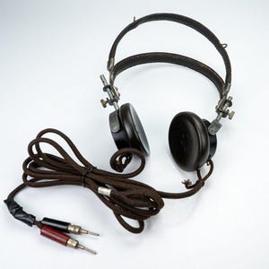 May include: Vintage black headphones with a brown cord and two silver-tipped plugs. The headphones have a leather headband and adjustable metal arms. The earcups are round and dark, with small holes. The cord is wrapped in a brown fabric.