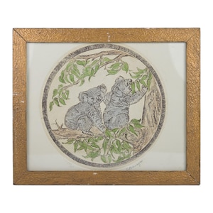 May include: Framed artwork featuring two koalas in a tree. The koalas are gray with detailed fur, surrounded by green leaves and branches. The artwork is in a circular design, framed in a gold-colored, textured frame.