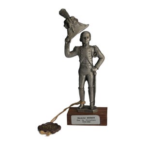 May include: A silver-colored figurine of a military figure holding a hat, mounted on a wooden base. The figurine is detailed with uniform features and a sword. The base has text that reads "Maréchal MONCEY Duc de Conegliano 1754-1842".