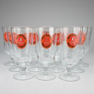 May include: A collection of clear glass beer goblets, each featuring a circular orange logo with the text Whitbread Imported English and a small horse graphic. The glasses have a stemmed base and a smooth, cylindrical bowl design.