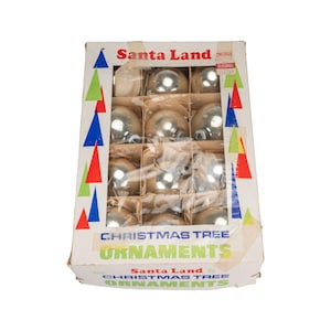 Pode incluir: Uma caixa vintage de enfeites para árvores de Natal. A caixa, rotulada como "Santa Land", contém vários enfeites de bolas de vidro prateadas. A caixa tem um design colorido com triângulos vermelhos, verdes e azuis. O texto "CHRISTMAS TREE ORNAMENTS" também é visível.