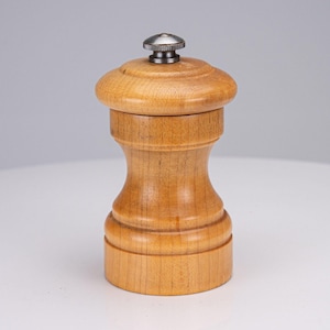 May include: A light brown wooden pepper mill with a smooth, hourglass-shaped body and a circular base. The top features a rounded wooden cap topped with a small, textured, dark metallic adjustment knob centred at the very peak of the device.