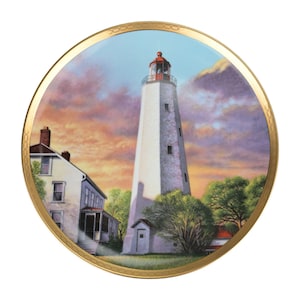 Könnte beinhalten: Dekorative Platte mit Leuchtturmmotiv. Der weiße Leuchtturm mit roter Spitze steht vor einem Sonnenuntergangshimmel. Ein Haus und Bäume sind ebenfalls abgebildet, alles innerhalb eines goldfarbenen Randes.