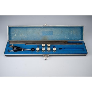 May include: An open, vintage metal case with a blue interior houses a set of precision measuring tools. The case contains a black and silver compass, several cylindrical measuring gauges, and a blue tube. The tools are arranged on a blue velvet-like surface.