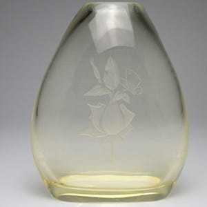May include: A clear, pale yellow glass vase with an oval, tapered silhouette. The front surface features a delicate, etched design of a single rose blossom, a rosebud, several leaves, and a small butterfly resting near the upper foliage.