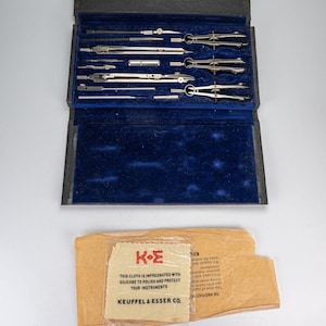 May include: A vintage drafting set in a black case with a blue velvet interior. The set includes various silver-colored compasses and tools. A paper packet with the text "KEUFFEL & ESSER CO." and "THIS CLOTH IS IMPREGNATED WITH SILICONE TO POLISH AND PROTECT YOUR INSTRUMENTS" is also present.