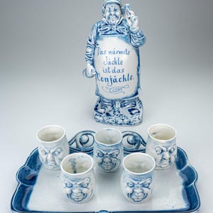 Può includere: Una bottiglia di liquore in ceramica blu e bianca a forma di chef, con bicchierini abbinati su un vassoio. La bottiglia ha il testo "Das wärmste Jäckle ist das Conjäckle."