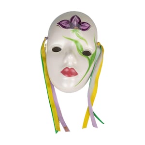 Puede incluir: Una máscara de cerámica blanca con rasgos pintados, incluyendo labios rojos, agujeros negros para los ojos y un diseño de flor púrpura en la frente. Cintas verdes y amarillas cuelgan de los lados. Un artículo decorativo único.