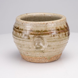 Puede incluir: Una pequeña taza de cerámica redonda con un esmalte moteado en tonos beige y bronceado. El exterior presenta estrías horizontales y una banda marrón más oscura cerca del borde, con una base rugosa de terracota sin esmaltar.