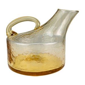 Puede incluir: Una jarra de vidrio transparente con una superficie texturizada y una base de color amarillo dorado. La jarra tiene un asa curva y un pico para verter. El vidrio tiene una textura martillada, agregando interés visual al diseño.