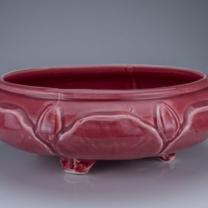 May include: A low, round, deep-red ceramic bowl featuring a glossy finish and a raised, molded floral petal pattern around the exterior. The bowl is supported by three small, integrated feet and has a wide, open top with a smooth, rounded rim.