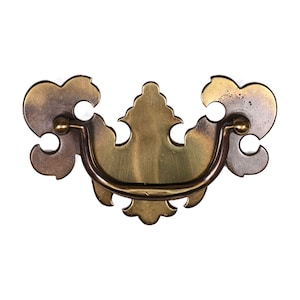 Puede incluir: Un tirador de cajón de latón de estilo vintage con un diseño decorativo. El tirador presenta un asa curva y detalles ornamentados y simétricos. El metal tiene una mezcla de tonos dorados y bronce, con una ligera pátina. El herraje está diseñado para muebles.