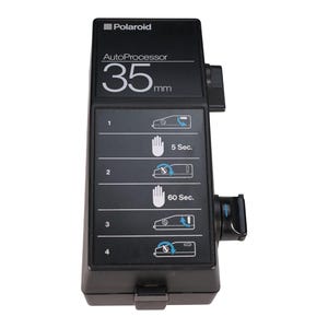 May include: A black Polaroid AutoProcessor for 35mm film. The device has the Polaroid logo at the top and instructions with hand symbols and time durations. It features a side knob and a bottom latch.