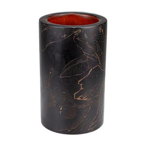 May include: A cylindrical, black container with a reddish-orange interior. The exterior features a detailed, etched design of leaves and foliage in a lighter brown color. The container is likely made of wood or a similar material.