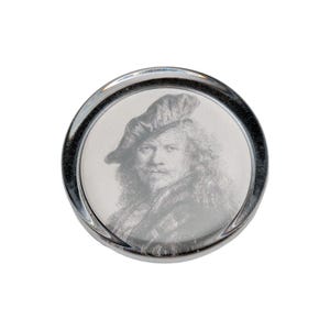 May include: A circular, metallic button featuring a detailed, monochromatic engraved portrait of a person wearing a beret. The button has a polished, reflective silver-toned rim that frames the central image, creating a distinct contrast.