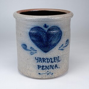 May include: A cylindrical stoneware crock with a speckled gray finish and a dark blue heart motif. The front features the blue text YARDLEY, PENNA. with decorative flourishes. The interior is dark, and the rim has a rounded, thick edge.