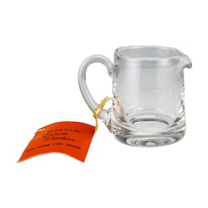 Può includere: Un boccale Lenox Spirits in vetro trasparente con un manico curvo e un beccuccio. Il boccale ha una base rettangolare e un'etichetta attaccata con un filo d'oro. L'etichetta è arancione con testo nero "Lenox Spirits Pitcher Hand-Blown Lead Crystal."