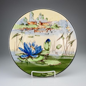 May include: A round ceramic plate featuring a hand-painted scene of a pond with a large, vibrant blue water lily in the foreground. The background depicts a stone wall, trees, and a calm water surface with green lily pads and tall aquatic plants.