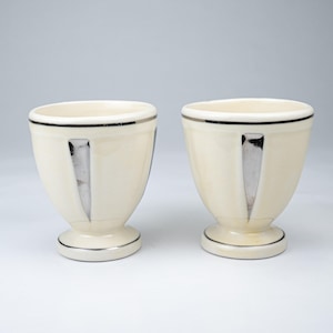 Peut inclure: Deux coquetiers en céramique de couleur crème présentant une forme de bol arrondie et lisse sur une base en piédestal. Chaque tasse est ornée d'un fin rebord argenté horizontal en haut et en bas, et d'une incrustation triangulaire argentée.