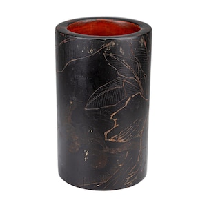 May include: A cylindrical, black container with a reddish-orange interior. The exterior features a detailed, etched design of leaves and other botanical elements in a lighter brown color. The container is likely made of wood or a similar material.