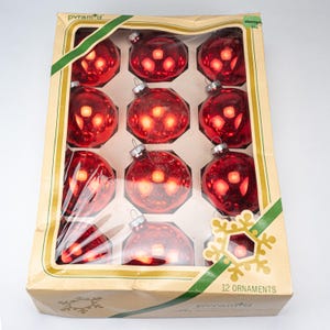 Puede incluir: Un juego de 12 adornos navideños de cristal rojo empaquetados en una caja de cartón rectangular con una ventana de plástico transparente. La caja muestra la marca Pyramid, un diseño de cinta verde y un copo de nieve con el texto 12 ORNAMENTS.