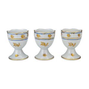 Puede incluir: Tres hueveras de porcelana blanca con ribetes dorados y diseños florales. Cada taza presenta una banda azul y detalles dorados. Las hueveras tienen una base de pedestal y un borde festoneado, creando un aspecto elegante y clásico.
