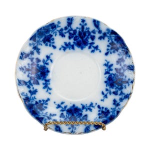May include: A white and blue floral patterned saucer with a gold rim. The blue floral design covers the saucer's surface. The saucer is displayed on a gold-colored metal stand. The saucer is a vintage item.