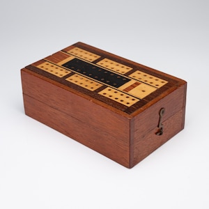 Könnte beinhalten: Eine rechteckige Cribbage-Spielbox aus Holz mit einer dunkel rötlich-braunen Holzaußenseite und einem Klappdeckel. Die Oberseite zeigt ein eingelegtes Muster aus hellen und dunklen Holzabschnitten mit mehreren Reihen von Löchern für Spielstifte.
