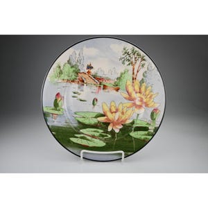 May include: A decorative plate featuring a serene water lily pond scene. The plate showcases yellow water lilies, lily pads, and a landscape with a bridge and trees. The plate has a black rim and is displayed on a white stand.