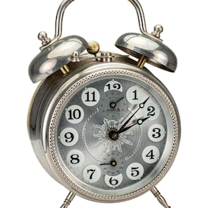 May include: A silver-toned, vintage alarm clock with a round face and two bells on top. The clock face is gray with black hands and white numerals. The clock has a handle and stands on four small legs.