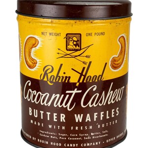 Puede incluir: Lata vintage de Robin Hood Coconut Cashew Butter Waffles. La lata cilíndrica tiene una tapa y base marrones con una etiqueta amarilla y marrón. La etiqueta presenta el texto "Coconut Cashew Butter Waffles" y "Made with Fresh Butter".