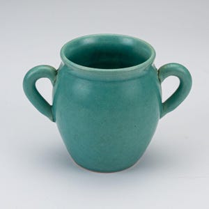 May include: A teal ceramic vessel with a rounded body and two symmetrical handles on opposite sides. The pot features a smooth, matte finish and a wide, open circular rim, showcasing a consistent solid color throughout the entire exterior surface.