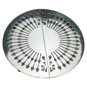 May include: A circular silver-toned metal trivet featuring a symmetrical design of radiating cut-out slots that resemble stylized flower stems. The object is composed of two semi-circular halves joined together, creating a decorative pattern.