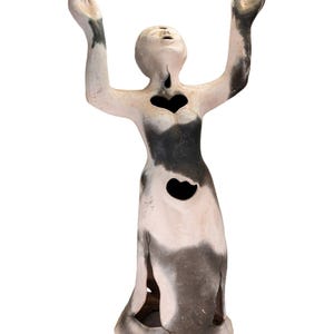May include: A ceramic sculpture of a figure with arms raised, featuring a heart-shaped cutout on the chest and abdomen. The figure is primarily white with black and gray accents, and the head is tilted upwards.