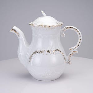 May include: A white porcelain teapot featuring a removable lid with a pointed finial. The teapot has a curved handle, a long spout, and embossed floral patterns on the body, all accented with worn gold-colored trim along the edges.